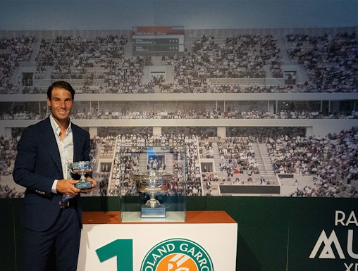 Man holding tennis trophies at Rafa Nadal Museum, Mallorca, with Roland Garros display in background.