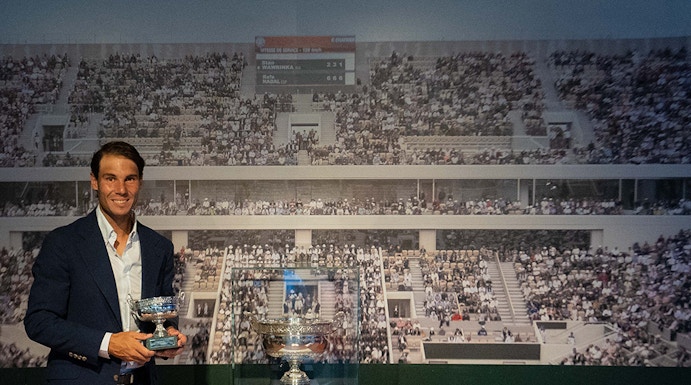 Man holding tennis trophies at Rafa Nadal Museum, Mallorca, with Roland Garros display in background.