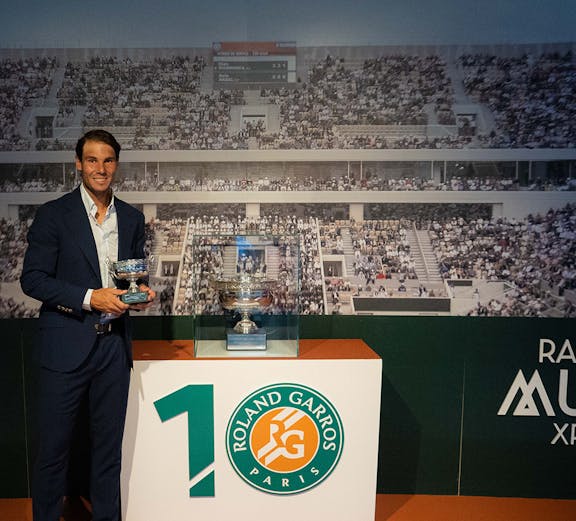 Man holding tennis trophies at Rafa Nadal Museum, Mallorca, with Roland Garros display in background.