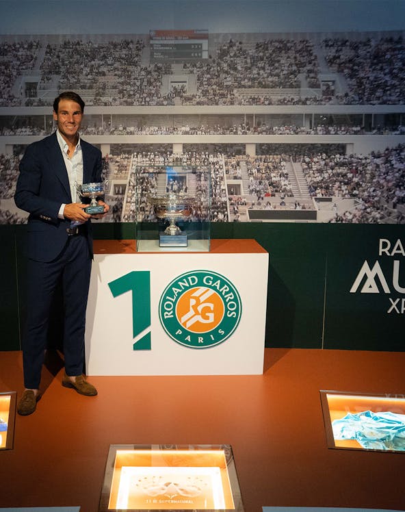 Man holding tennis trophies at Rafa Nadal Museum, Mallorca, with Roland Garros display in background.