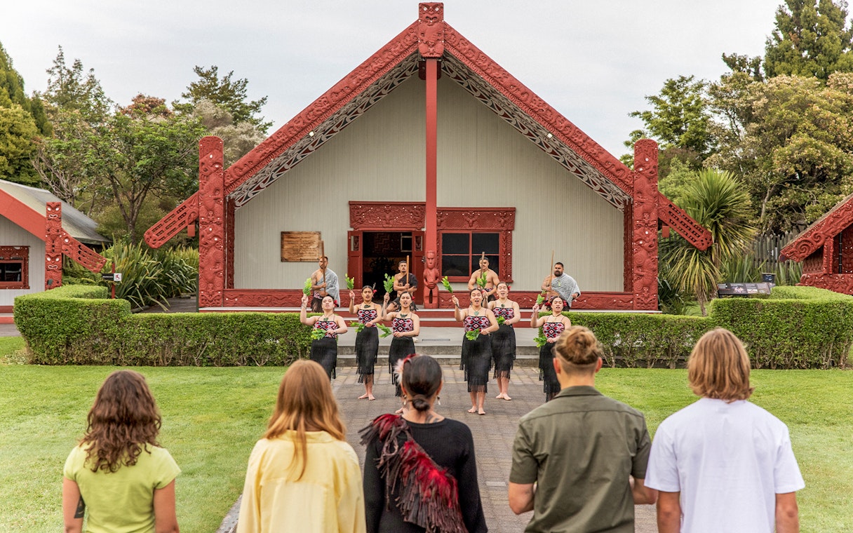 Maori cultural performance at Te Puia, New Zealand, with traditional meeting house in background.