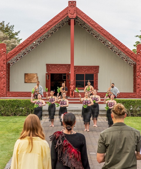 Maori cultural performance at Te Puia, New Zealand, with traditional meeting house in background.