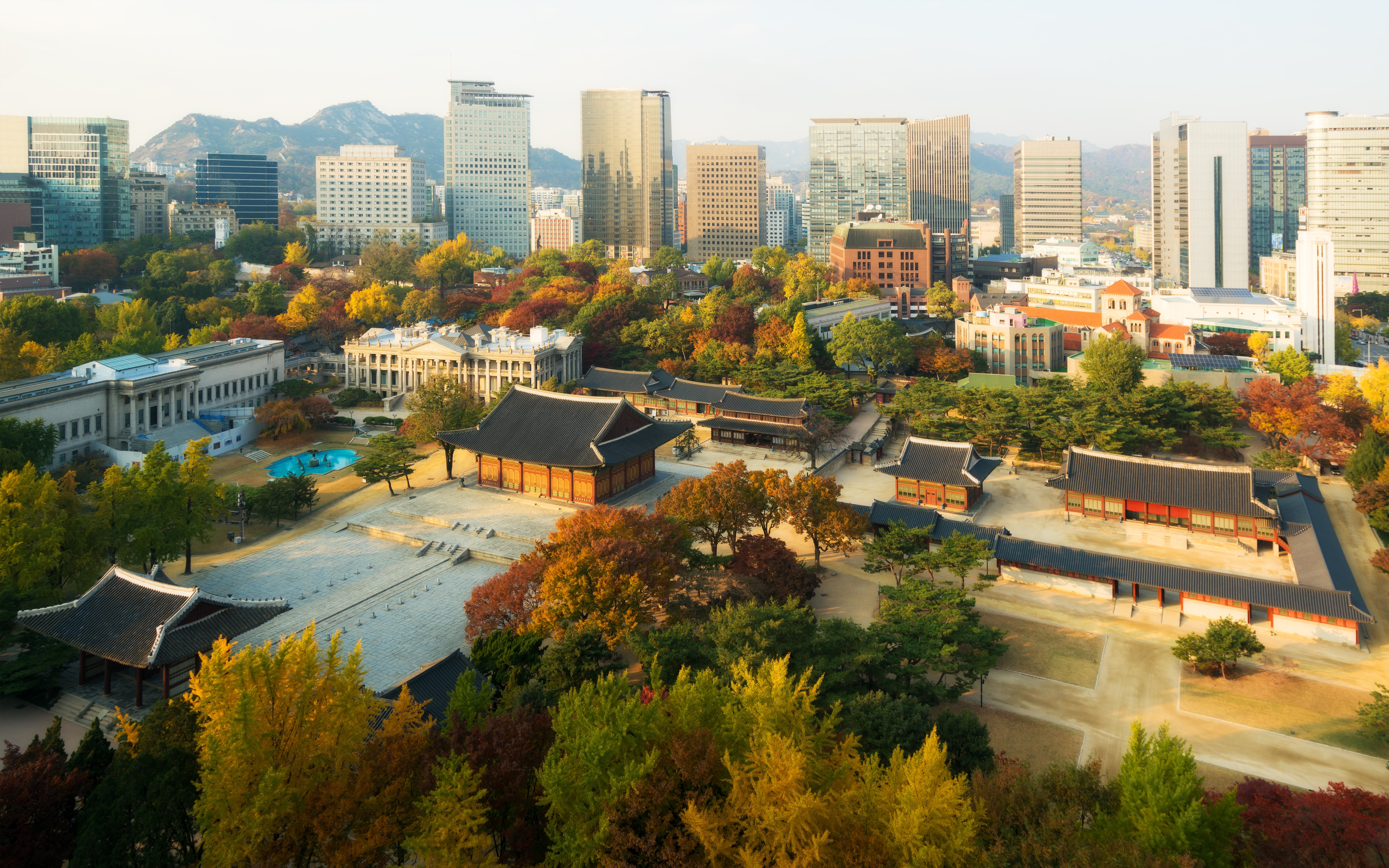 Deoksugung Palace surrounded by autumn foliage with Seoul city skyline in the background.
