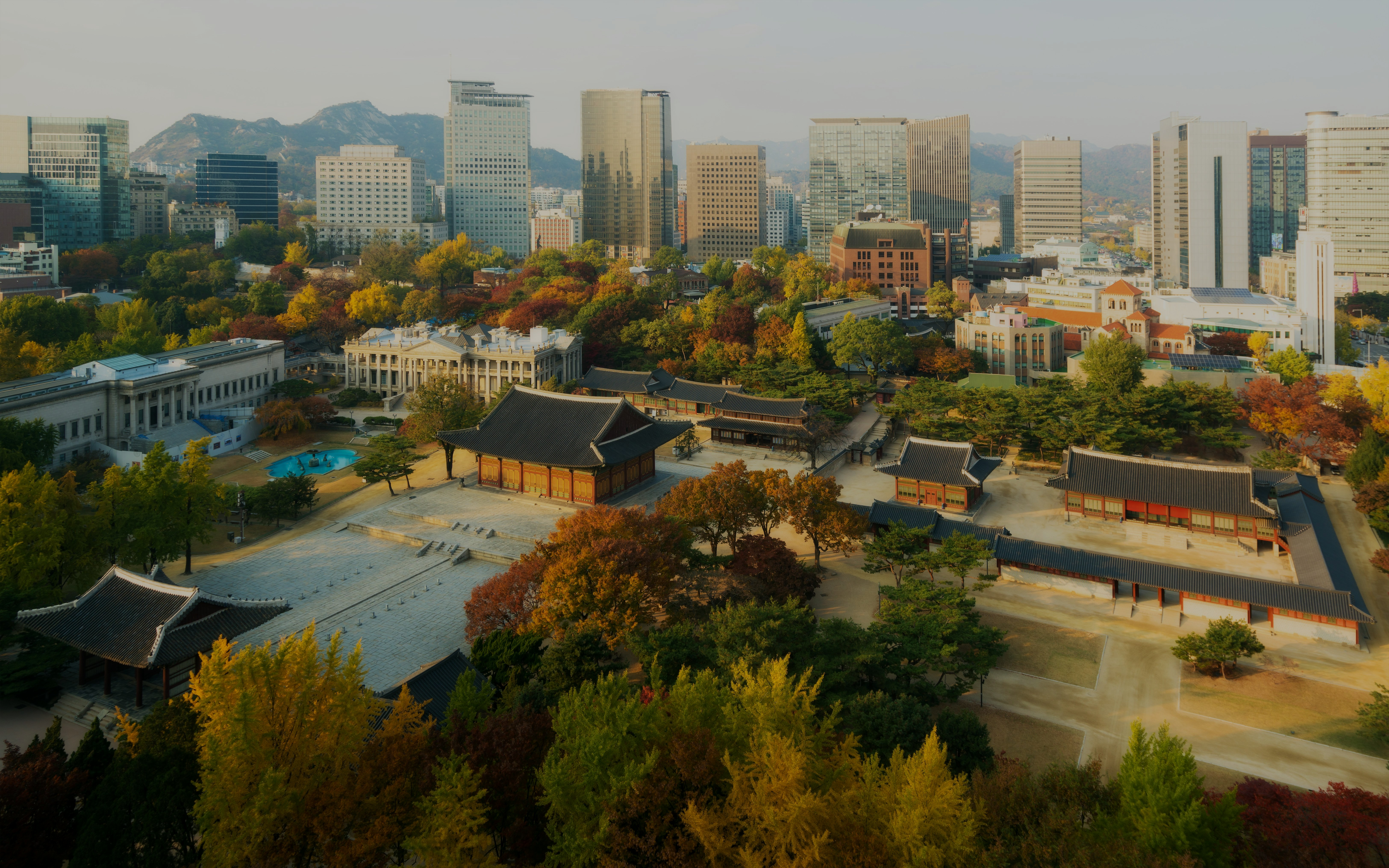 Deoksugung Palace surrounded by autumn foliage with Seoul city skyline in the background.