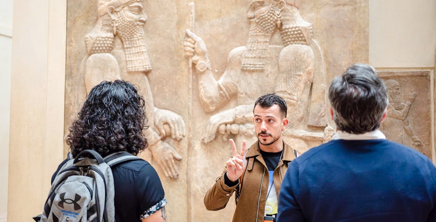 Guide explaining Assyrian relief to tourists at Louvre Museum, France.