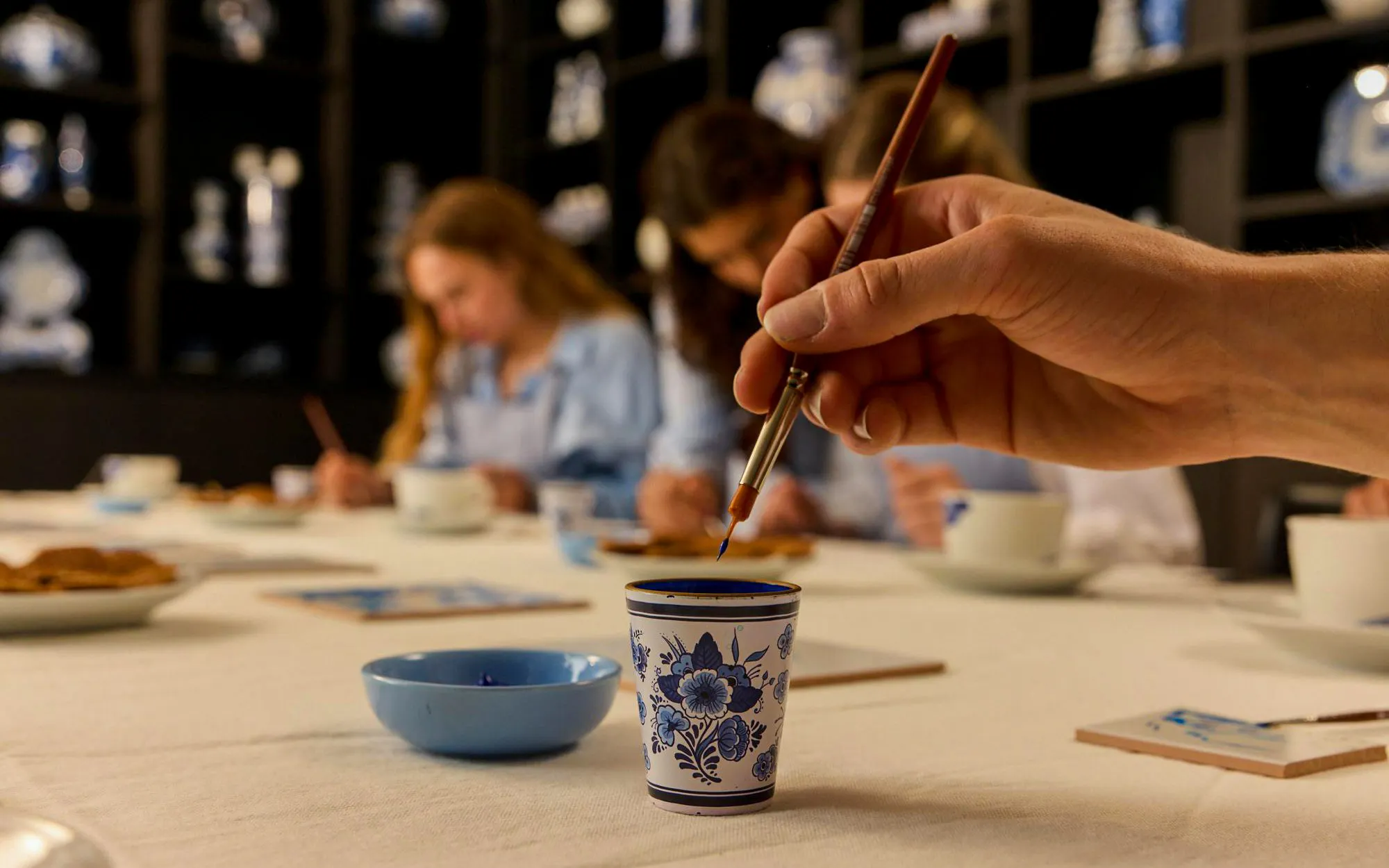 Hand painting a traditional Dutch blue tile with guests in the background at a workshop.