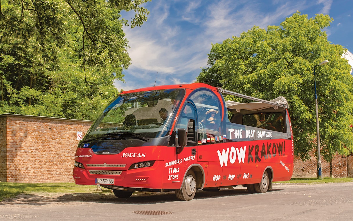 Red Kraków hop-on hop-off tour bus parked near a brick wall and trees.