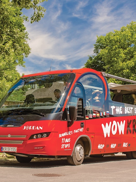 Red Kraków hop-on hop-off tour bus parked near a brick wall and trees.