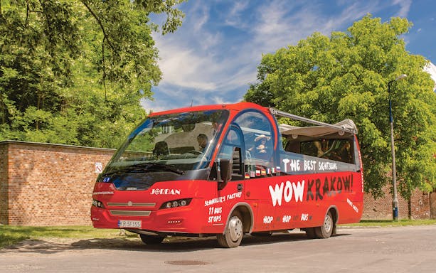 Red Kraków hop-on hop-off tour bus parked near a brick wall and trees.