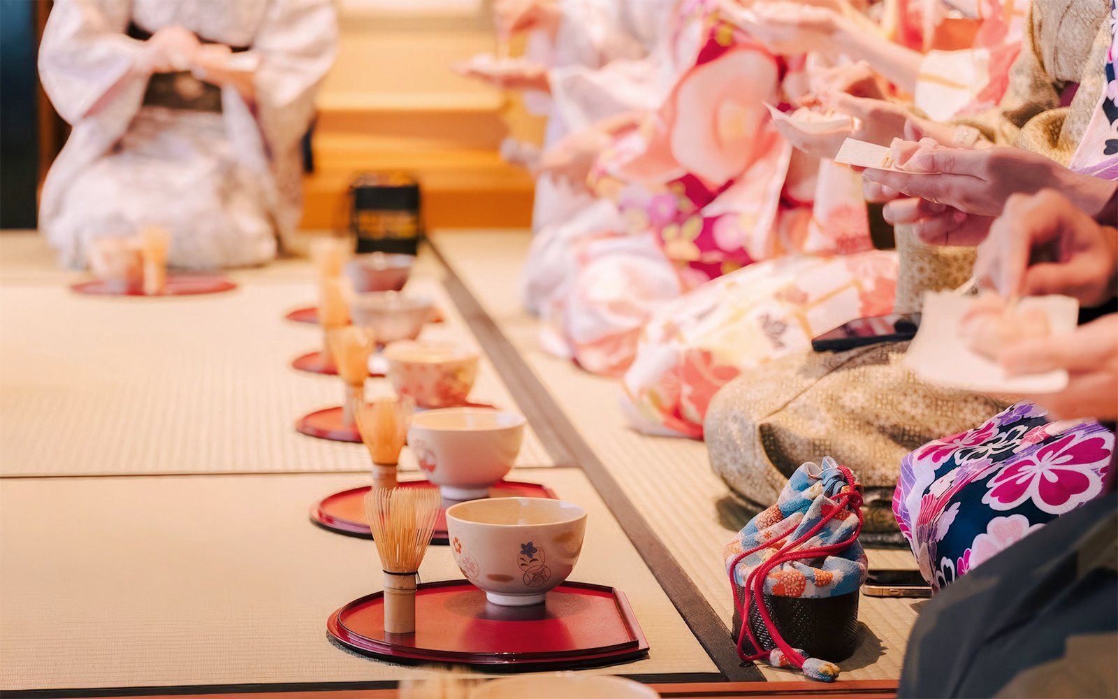 Tea ceremony in progress with participants in traditional attire, bowls, and tea tools on tatami mats.