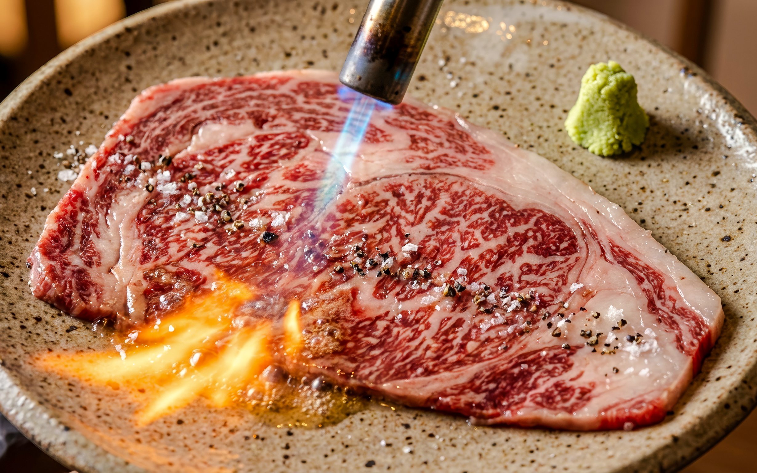 Wagyu beef being seared with a torch, garnished with salt and pepper, wasabi on the side.