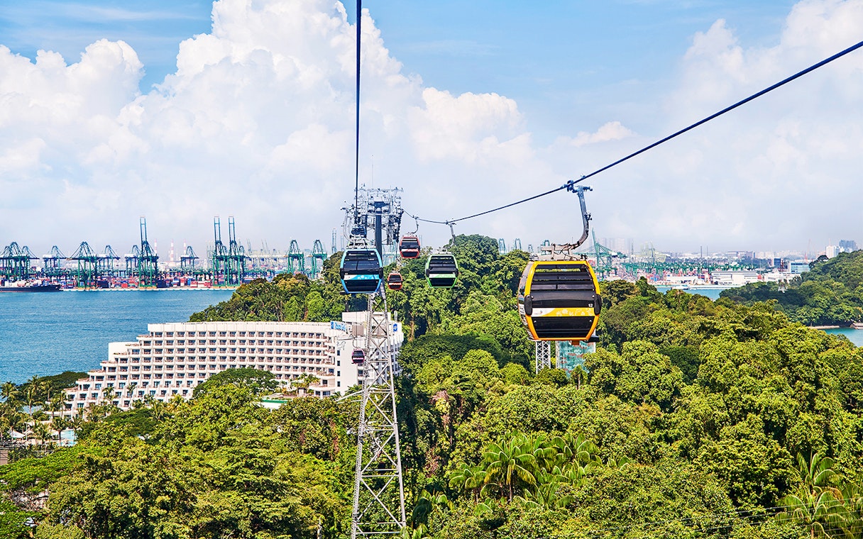 Cable cars over Sentosa Island with view of lush greenery and Singapore harbor.