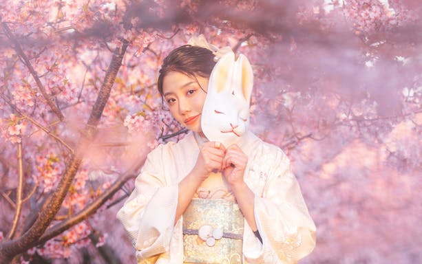 Kimono-clad person with fox mask under cherry blossoms at Kiyomizu Dera, Kyoto.