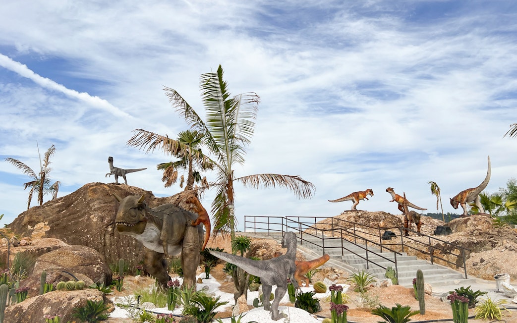 Dinosaurs in a desert landscape with palm trees and rocky terrain, Dino Desert Ticket.