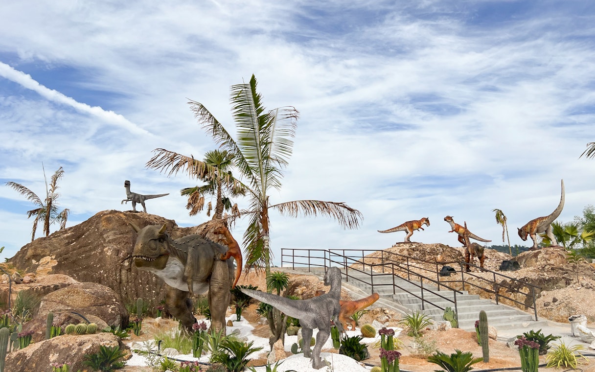 Dinosaurs in a desert landscape with palm trees and rocky terrain, Dino Desert Ticket.