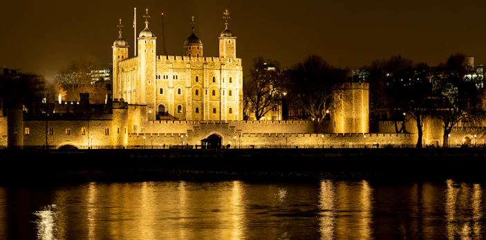 Tower of London illuminated at night, reflecting on the Thames River.