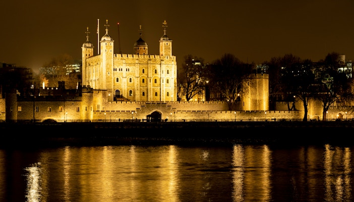 Tower of London illuminated at night, reflecting on the Thames River.