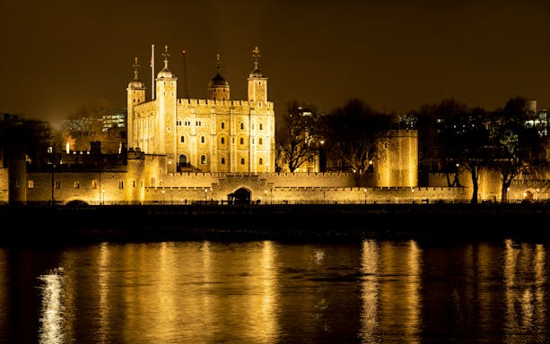 Tower of London illuminated at night, reflecting on the Thames River.