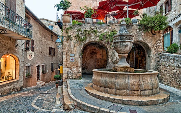 Stone fountain in a cobblestone street of Saint Paul de Vence, France, with rustic buildings and red umbrellas.