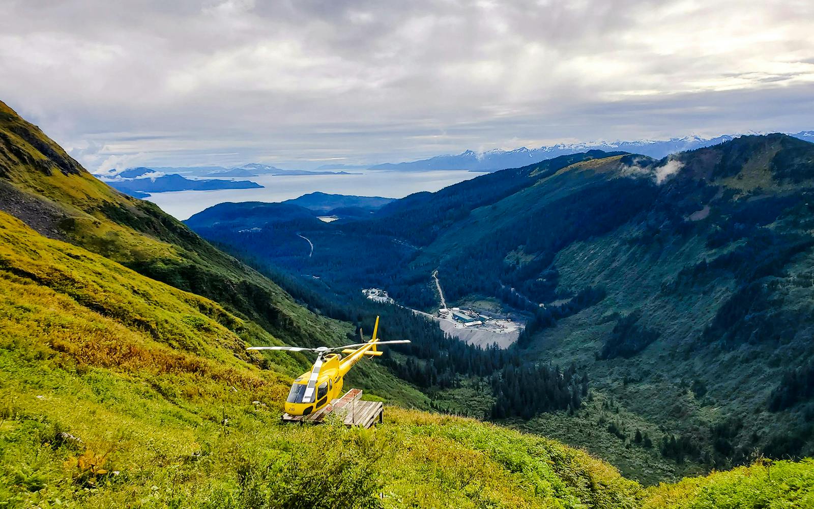 Yellow helicopter on a lush mountain slope in Juneau with distant glacier views.