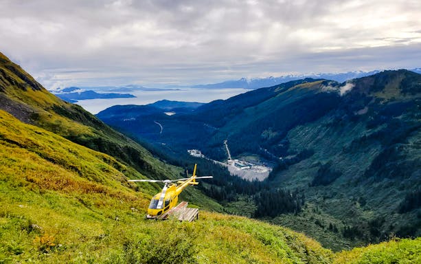 Yellow helicopter on a lush mountain slope in Juneau with distant glacier views.