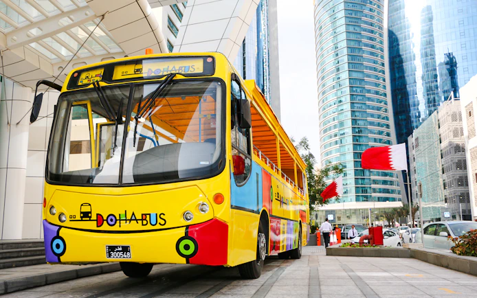 Colorful Doha tour bus in front of modern skyscrapers with Qatar flags.