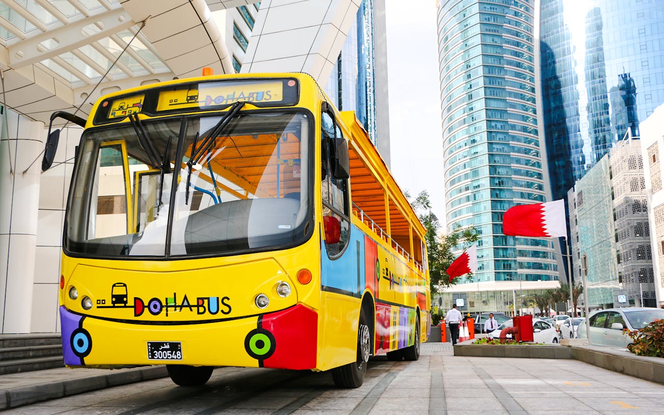 Colorful Doha tour bus in front of modern skyscrapers with Qatar flags.
