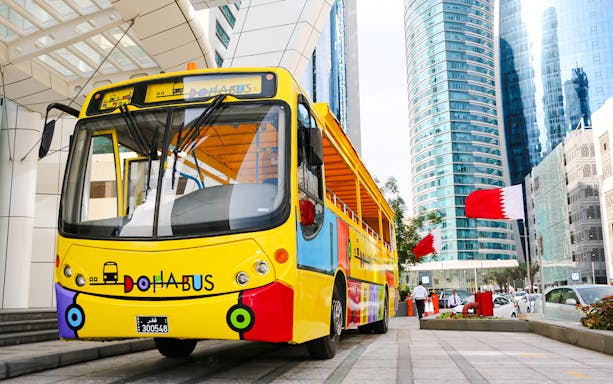 Colorful Doha tour bus in front of modern skyscrapers with Qatar flags.