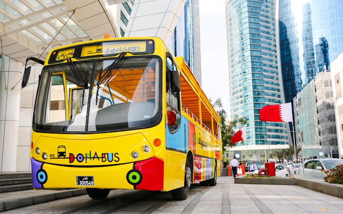 Colorful Doha tour bus in front of modern skyscrapers with Qatar flags.