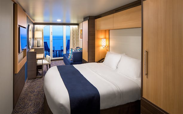 Cruise ship room with bed, desk, and balcony view of the ocean.