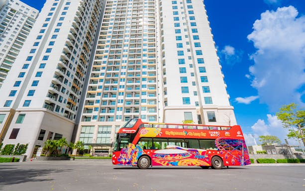 Red double-decker bus for Ha Long Bay hop-on hop-off tour parked near tall buildings.