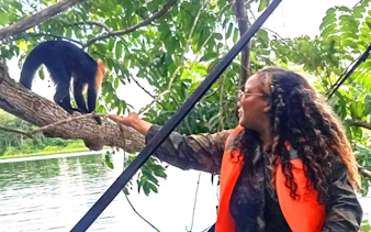 Person interacting with a black howler monkey on a tree branch by a river.