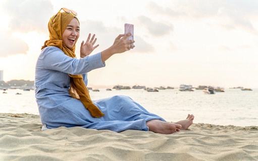 Woman taking a selfie on a sandy beach in Malaysia with boats in the background.