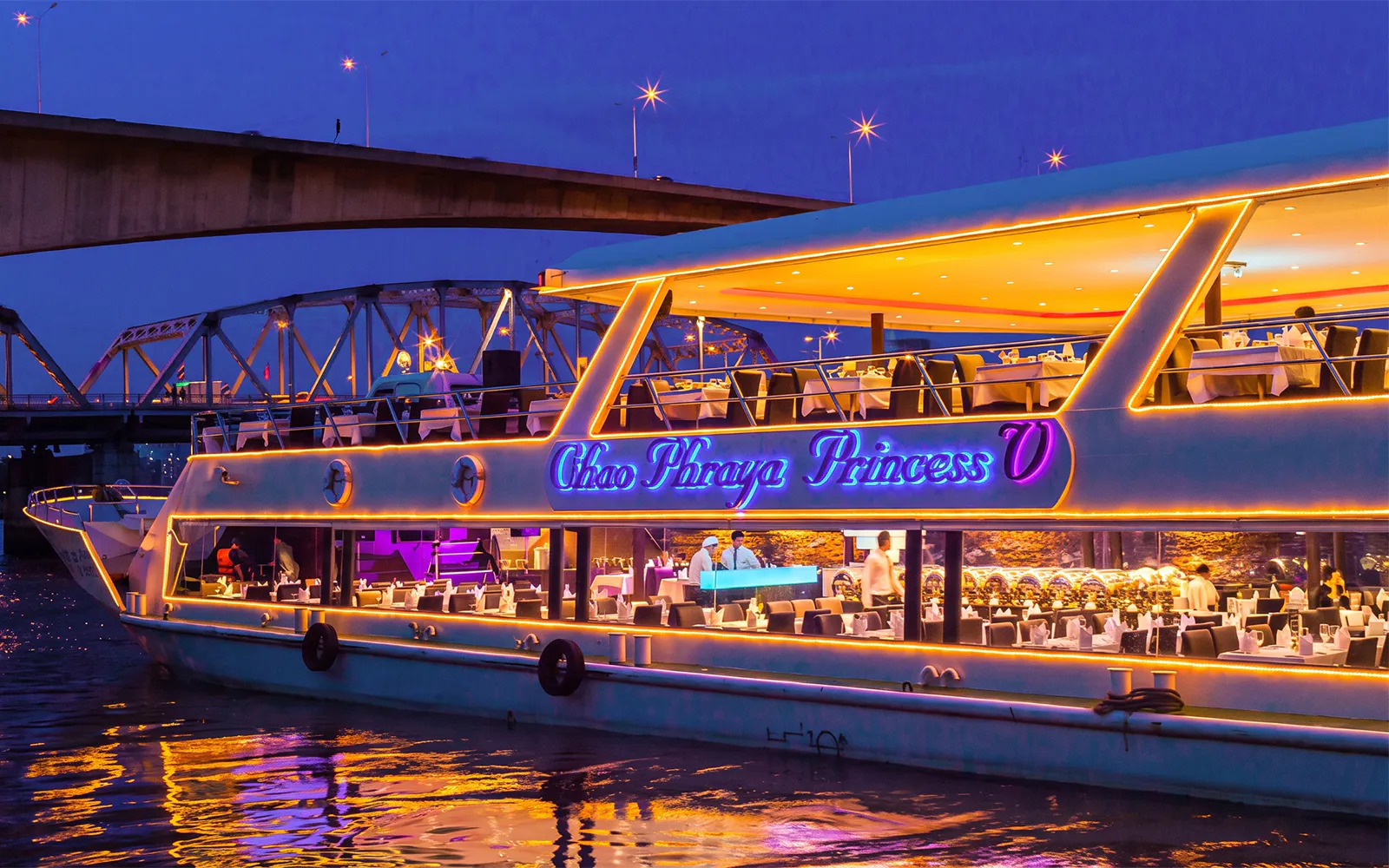 Chao Phraya Princess cruise lit up at night under a bridge, tables set for New Year's Eve dinner.