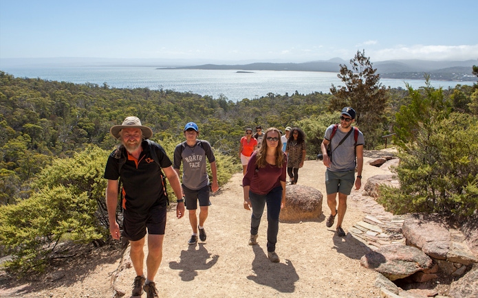 Group hiking on a trail with views of Wineglass Bay, Tasmania in the background.