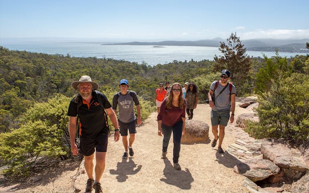 Group hiking on a trail with views of Wineglass Bay, Tasmania in the background.