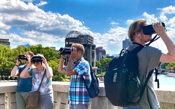 Visitors using VR headsets at Hiroshima Peace Park with Atomic Bomb Dome in background.