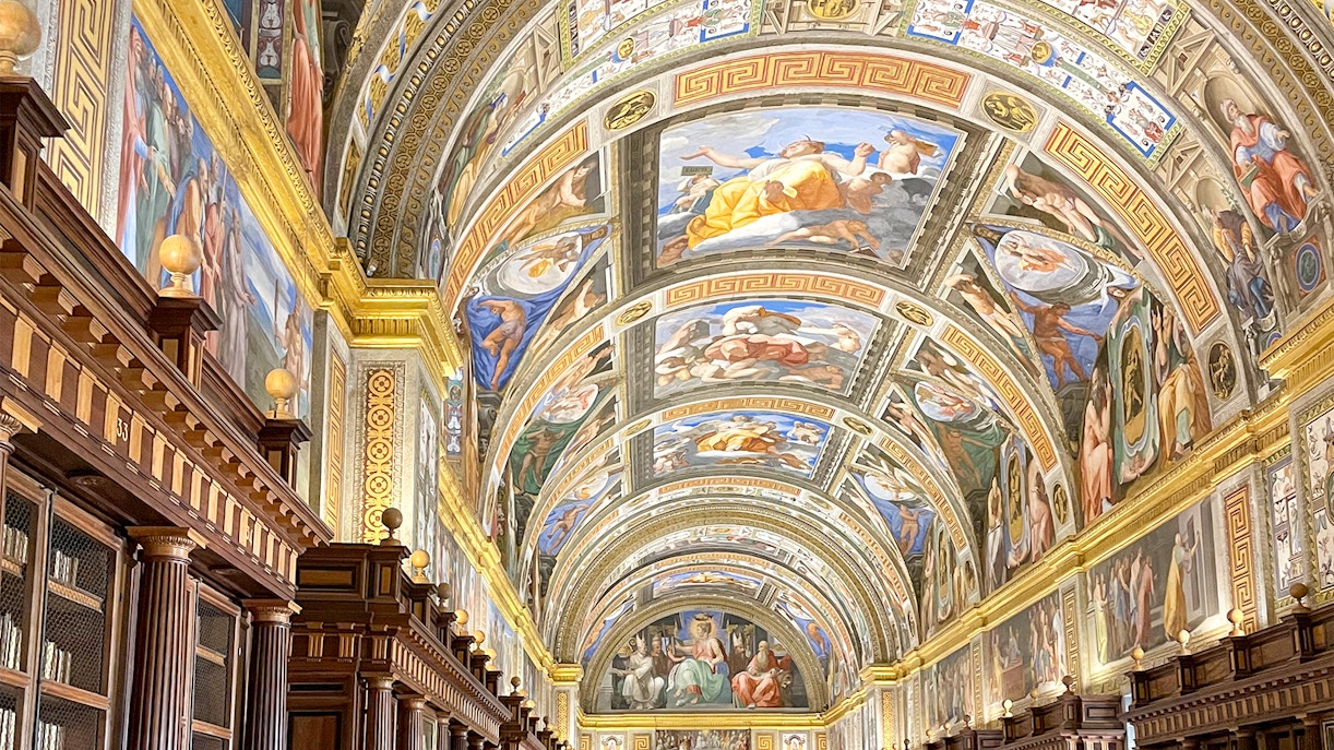 El Escorial Library interior with ornate ceiling frescoes and rows of ancient books in Madrid, Spain.