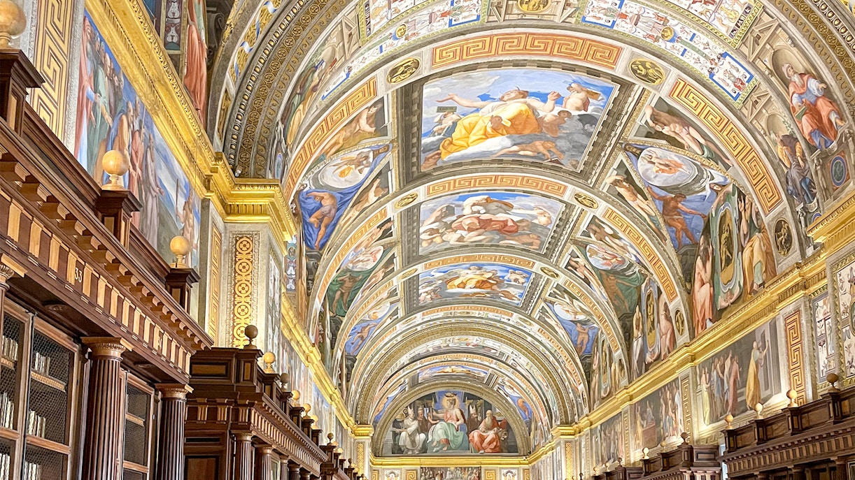 El Escorial Library interior with ornate ceiling frescoes and rows of ancient books in Madrid, Spain.