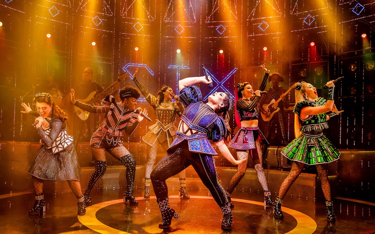 Performers in vibrant costumes singing on stage during the musical "Six.