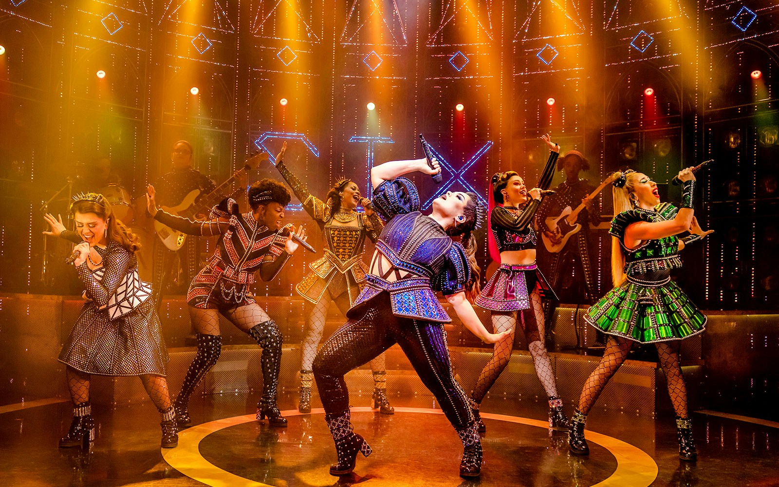 Performers in vibrant costumes singing on stage during the musical "Six.