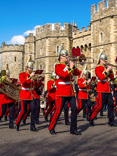 Guards in red uniforms marching with instruments in front of Windsor Castle.
