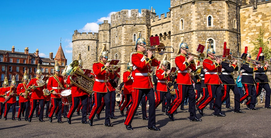 Guards in red uniforms marching with instruments in front of Windsor Castle.