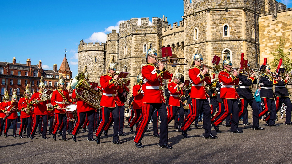Guards in red uniforms marching with instruments in front of Windsor Castle.