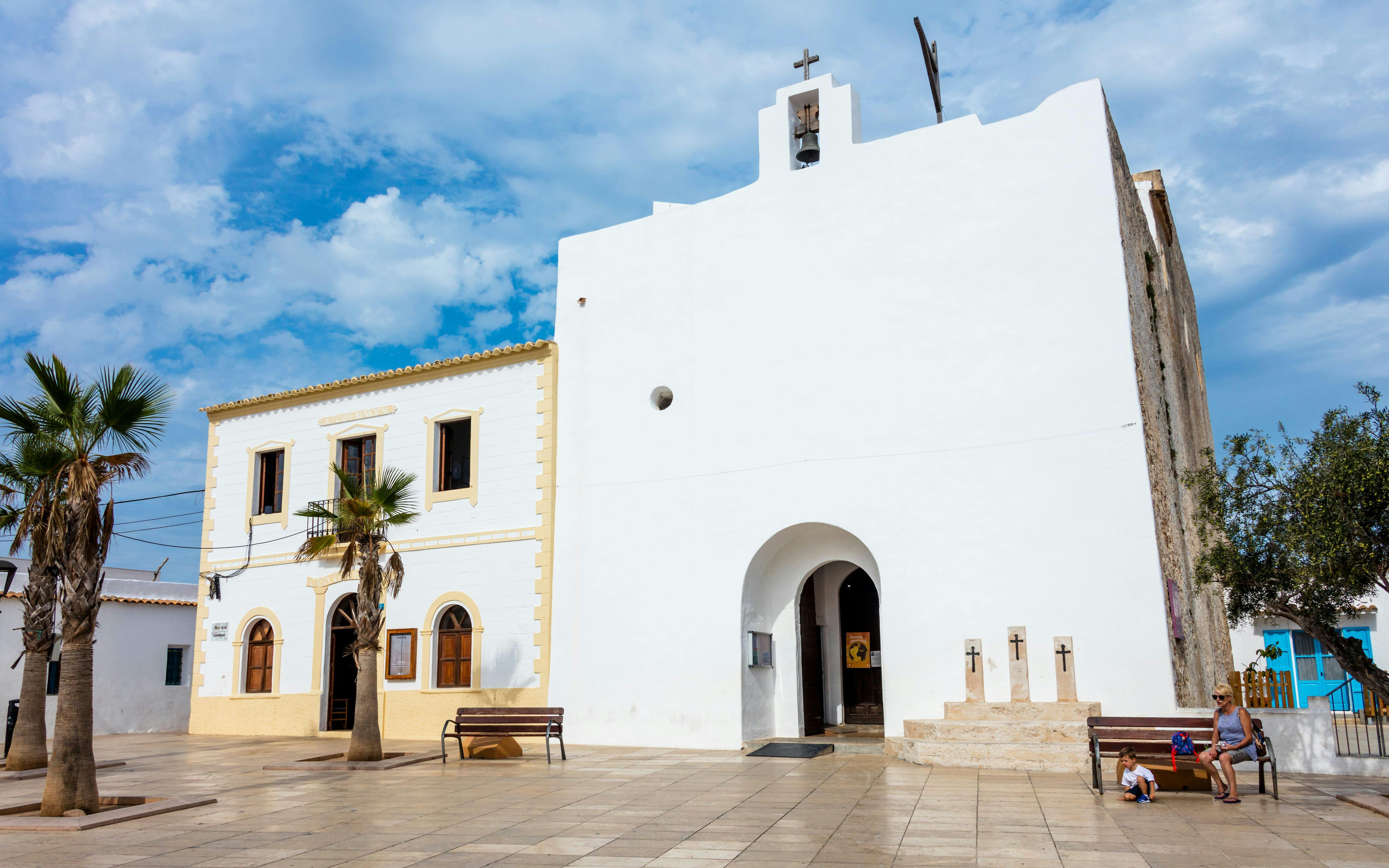 Church of Sant Francesc Xavier in Formentera's main square, Spain, with people sitting nearby.