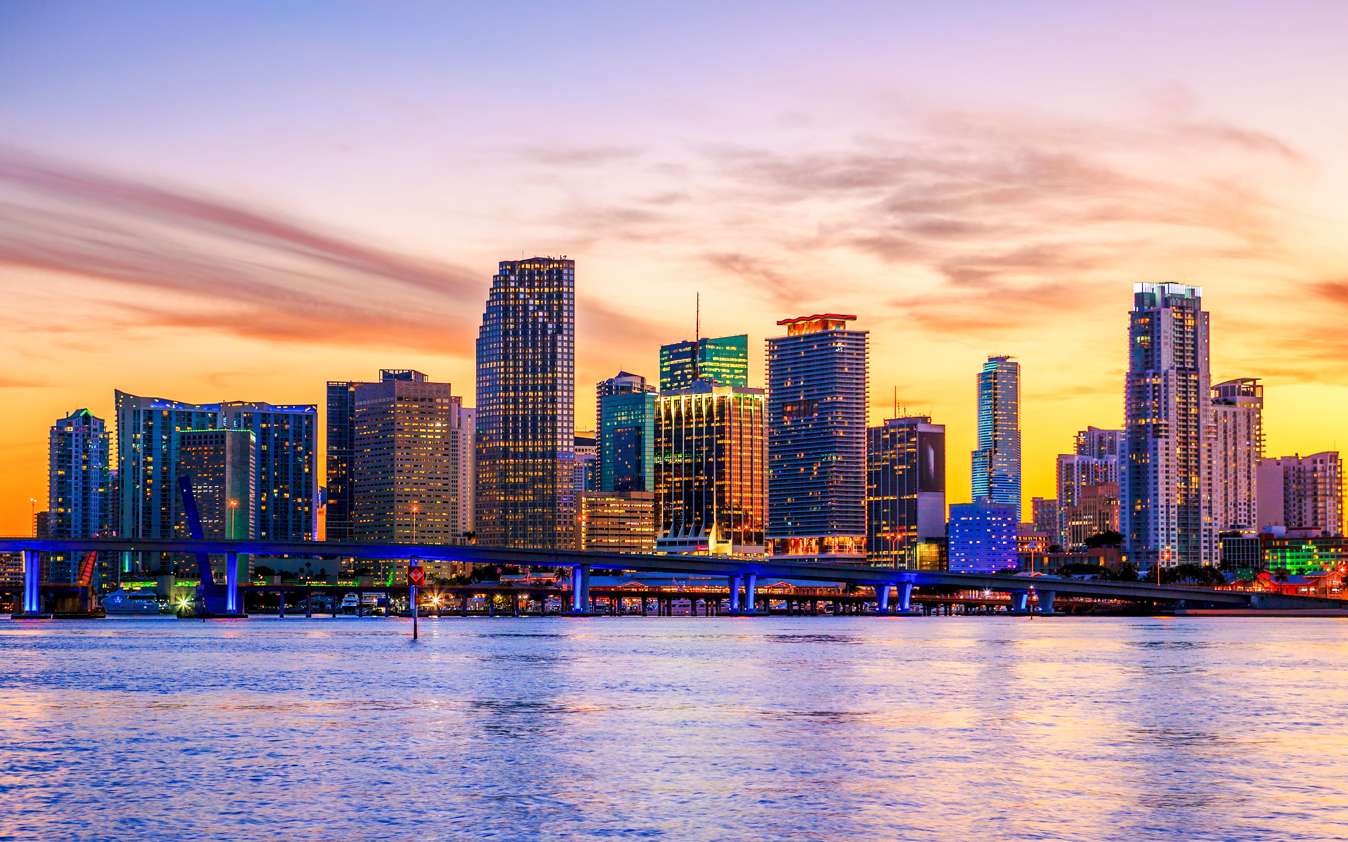Miami skyline at sunset with illuminated buildings and a bridge over the water.