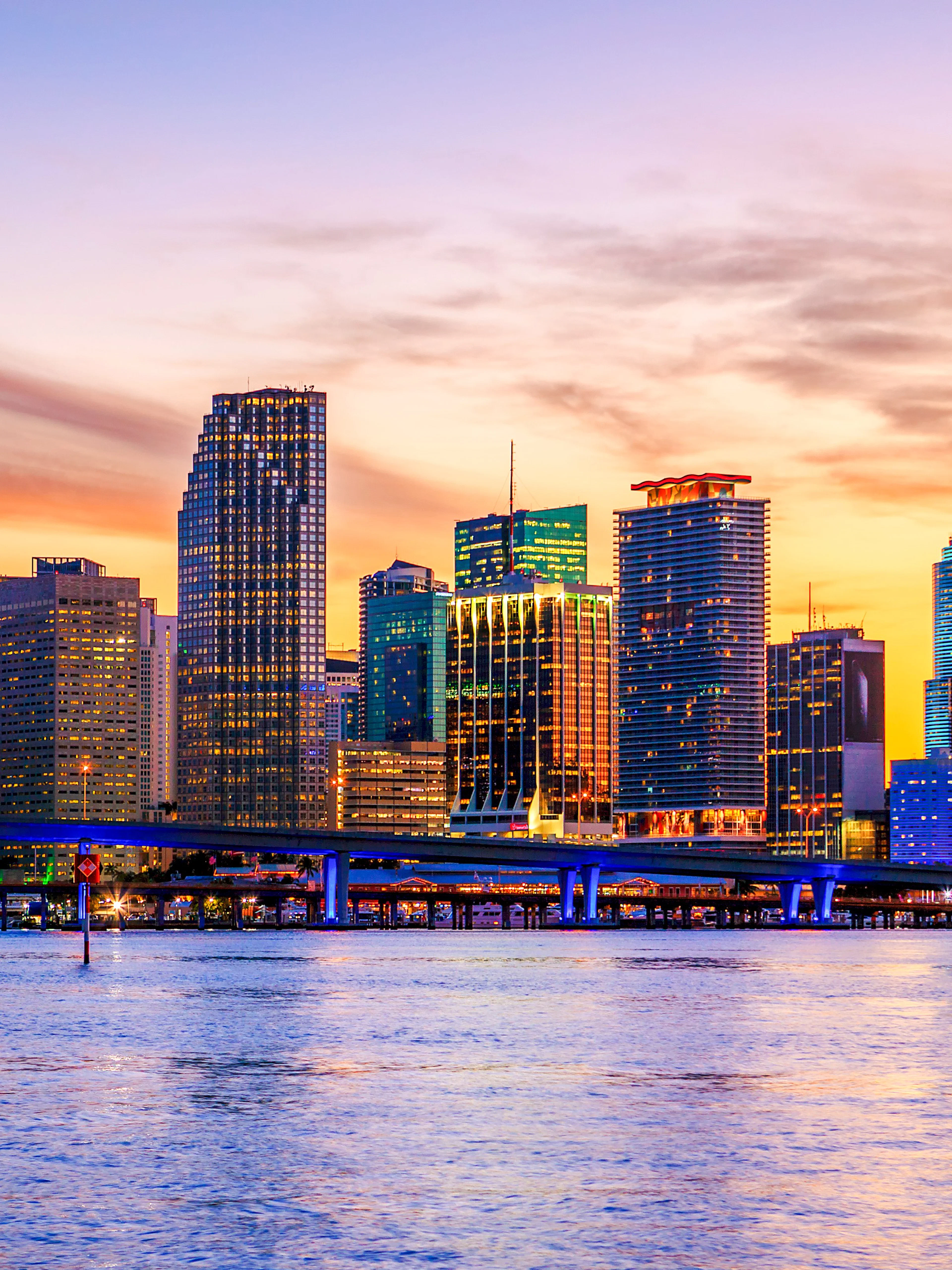 Miami skyline at sunset with illuminated buildings and a bridge over the water.