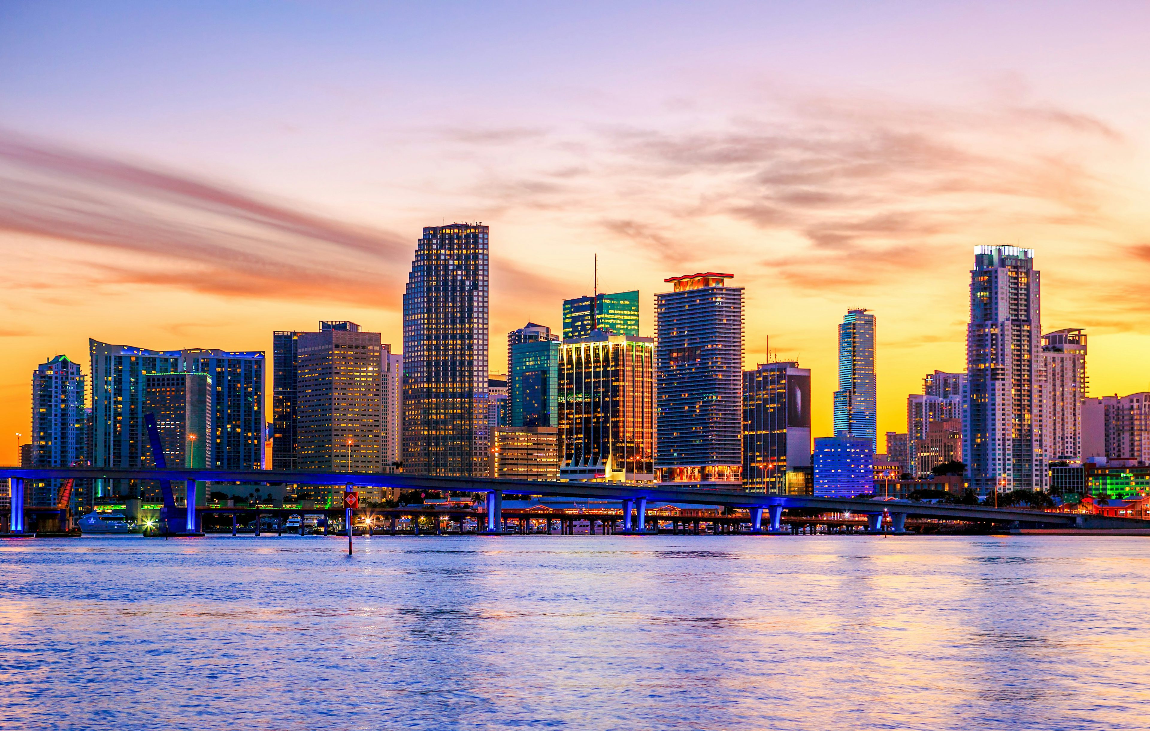 Miami skyline at sunset with illuminated buildings and a bridge over the water.