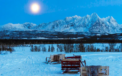 Sleds on snowy landscape under moonlit sky in Tromso with mountain backdrop.