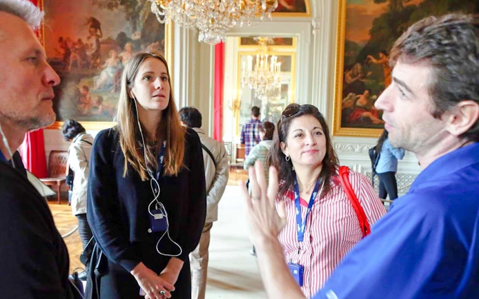 Tour group listening to a guide in a Versailles palace room with ornate paintings and chandeliers.